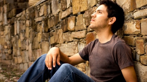 Um jovem sentado e encostado em um muro de pedra.