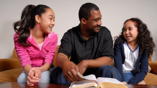 Um pai e suas duas filhas conversando diante de uma Bíblia aberta.