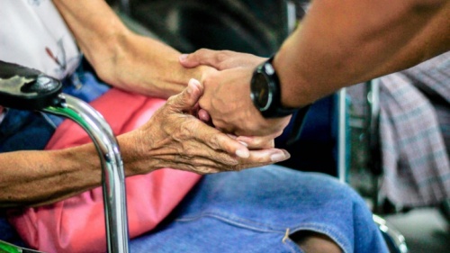 Um homem segurando a mão de uma mulher idosa sentada em uma cadeira de rodas.