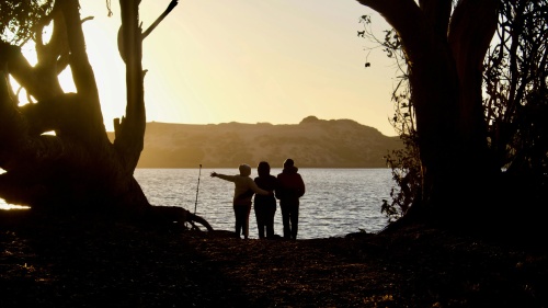 Três pessoas à beira de um lago admirando o brilho do sol.