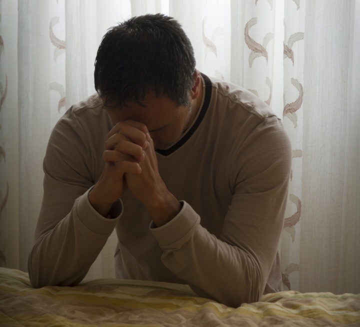 Um homem orando ao lado de uma cama.