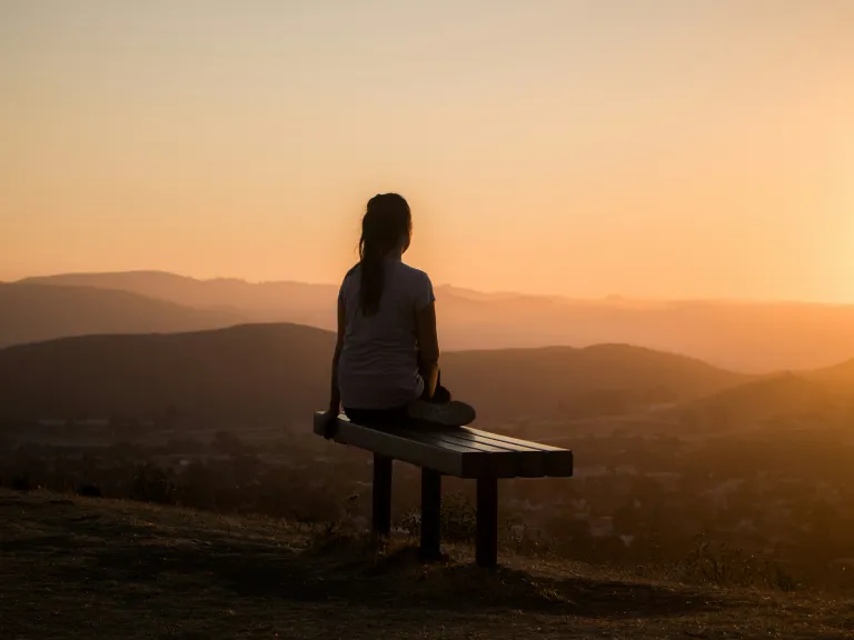 A woman sitting on a bench looking out at sunset vista.
