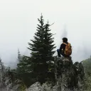 Um caminhante solitário com uma mochila senta-se em uma saliência rochosa com vista para uma floresta de coníferas cheia de neblina.