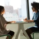 Duas mulheres que conversam sentadas ao redor de uma mesa.