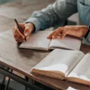 Um homem escrevendo num caderno ao lado de uma Bíblia aberta.