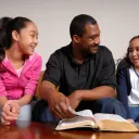 Um pai e suas duas filhas conversando diante de uma Bíblia aberta.